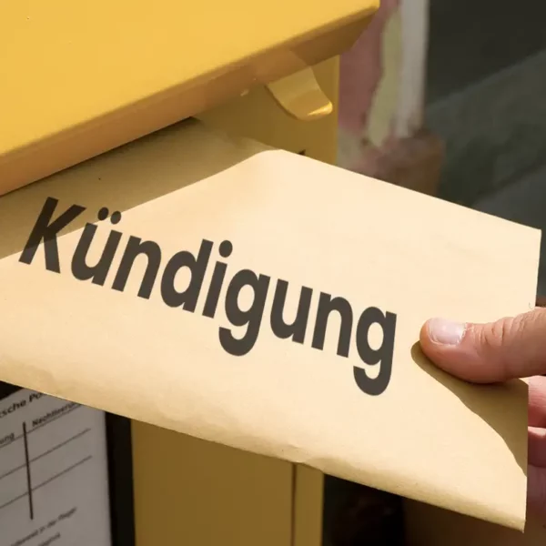 Eine Hand steckt einen braunen Umschlag mit dem fettgedruckten Wort „Kündigung“ in einen gelben Briefkasten der Deutschen Post.