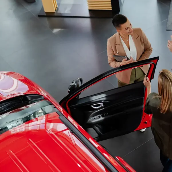 Eine Verkäuferin im Autohaus berät ein Paar neben einem leuchtend roten Auto mit geöffneter Fahrertür.