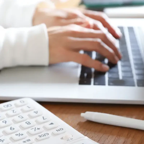Hände tippen auf der Tastatur eines Laptops, davor liegen ein weißer Taschenrechner und ein Stift auf einem Holztisch.