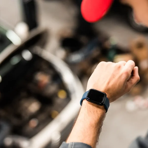 Ein Kfz-Mechatroniker mit Kaffeebecher blickt in einer Werkstatt auf seine Smartwatch; im unscharfen Hintergrund steht ein Auto mit geöffneter Motorhaube.