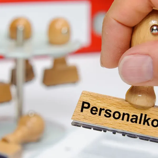 Alternativtext: Eine Hand drückt einen Holzstempel mit der Aufschrift „Personalkosten“ auf ein Papierfeld, im unscharfen Hintergrund stehen weitere Stempel in einem Karussell vor einem roten Aktenordner.