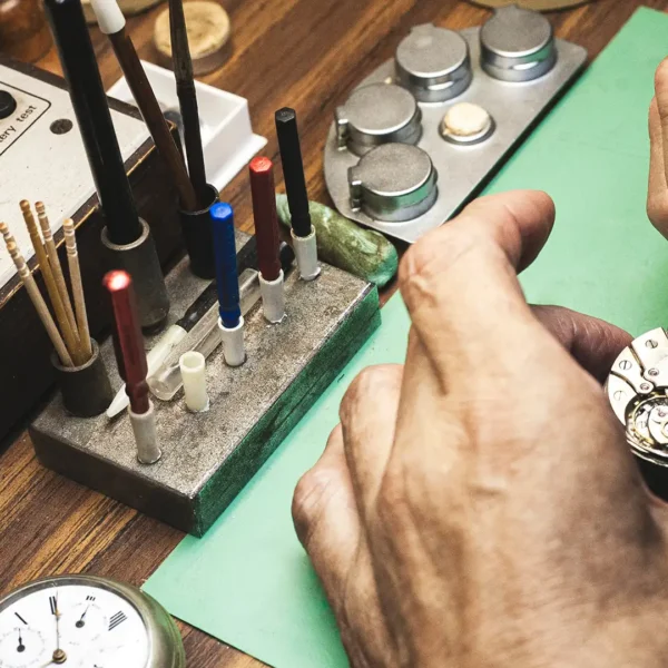 Nahaufnahme der Hände eines Uhrmachers, der mit einer Pinzette das mechanische Uhrwerk einer Taschenuhr an einem Arbeitstisch repariert.