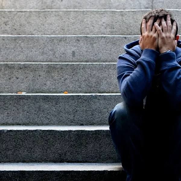 Ein Mann im blauen Kapuzenpullover sitzt zusammengesunken auf einer Steintreppe und verbirgt sein Gesicht in den Händen.