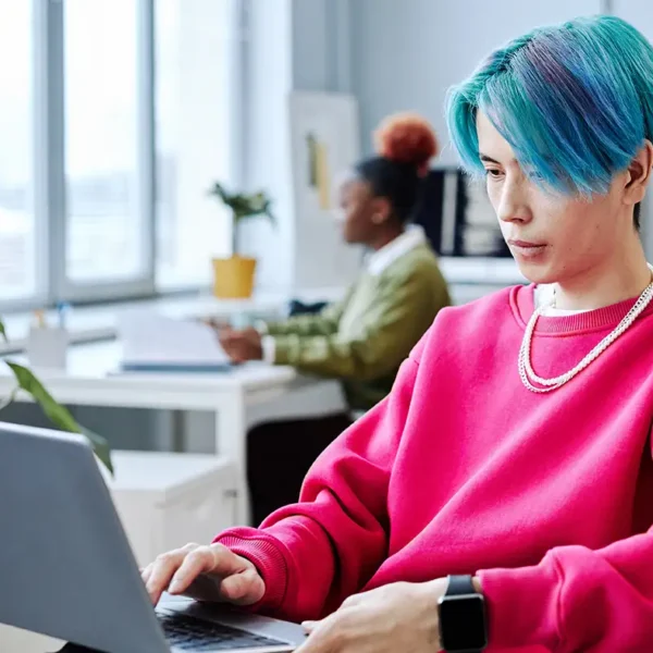 Eine junge Person mit blauen Haaren und pinkem Pullover arbeitet fokussiert an einem Laptop in einem hellen Büro.